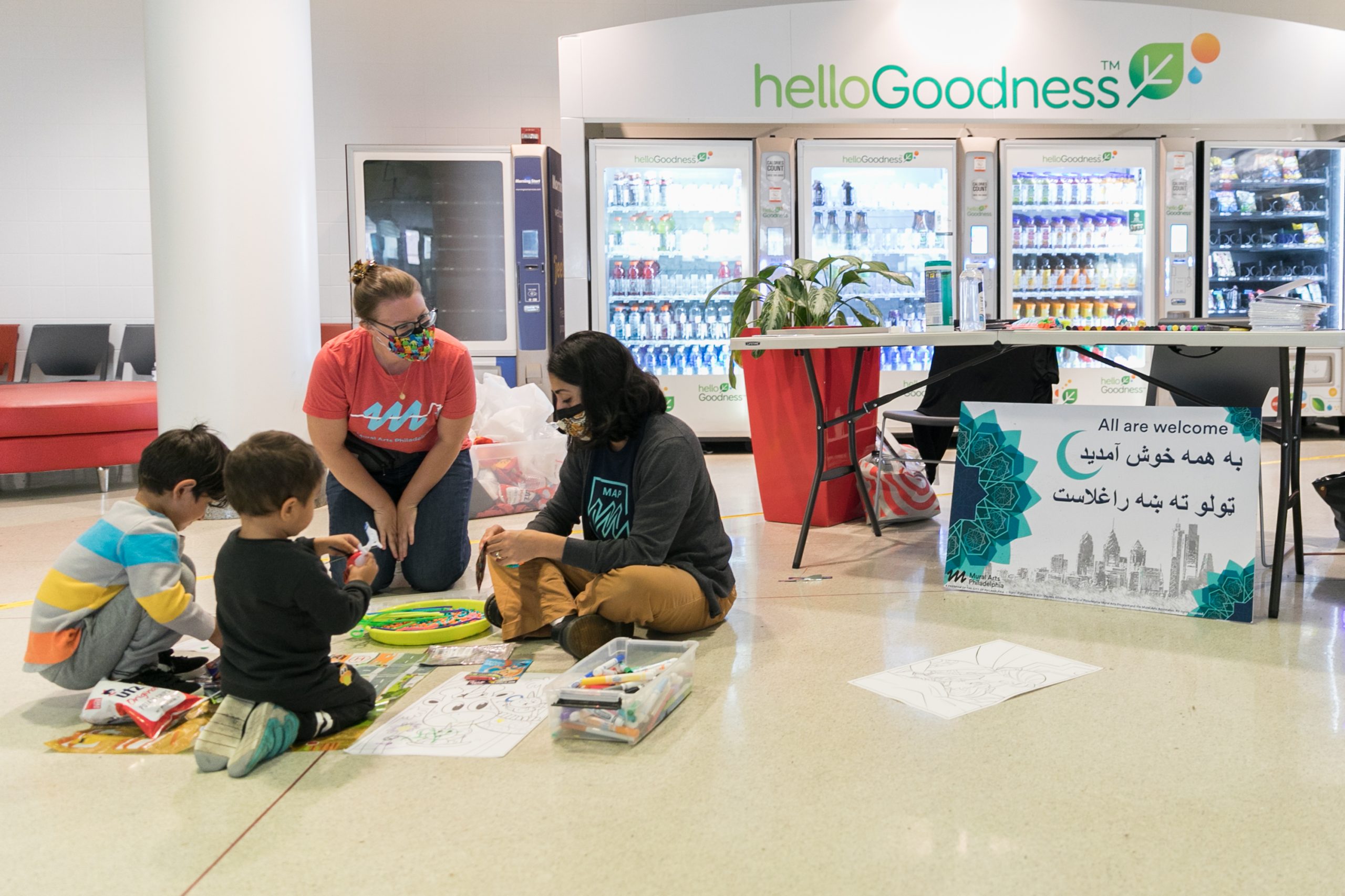Two Porch Light staff members sit on the floor with two young children, surrounded by papers and markers and a sign that says All Are Welcome in English, Pashto and Farsi at the airport.