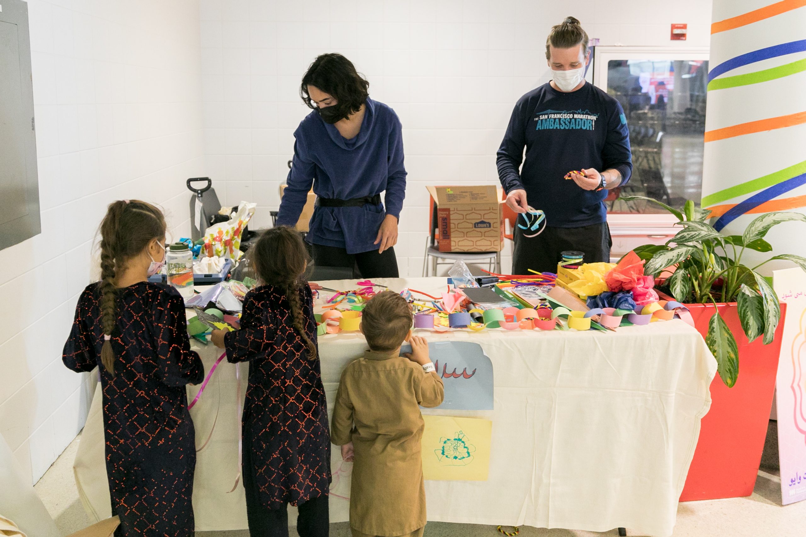 Two volunteers stand behind a table full of crafts in a corner of the airport, interacting with three children of various ages.