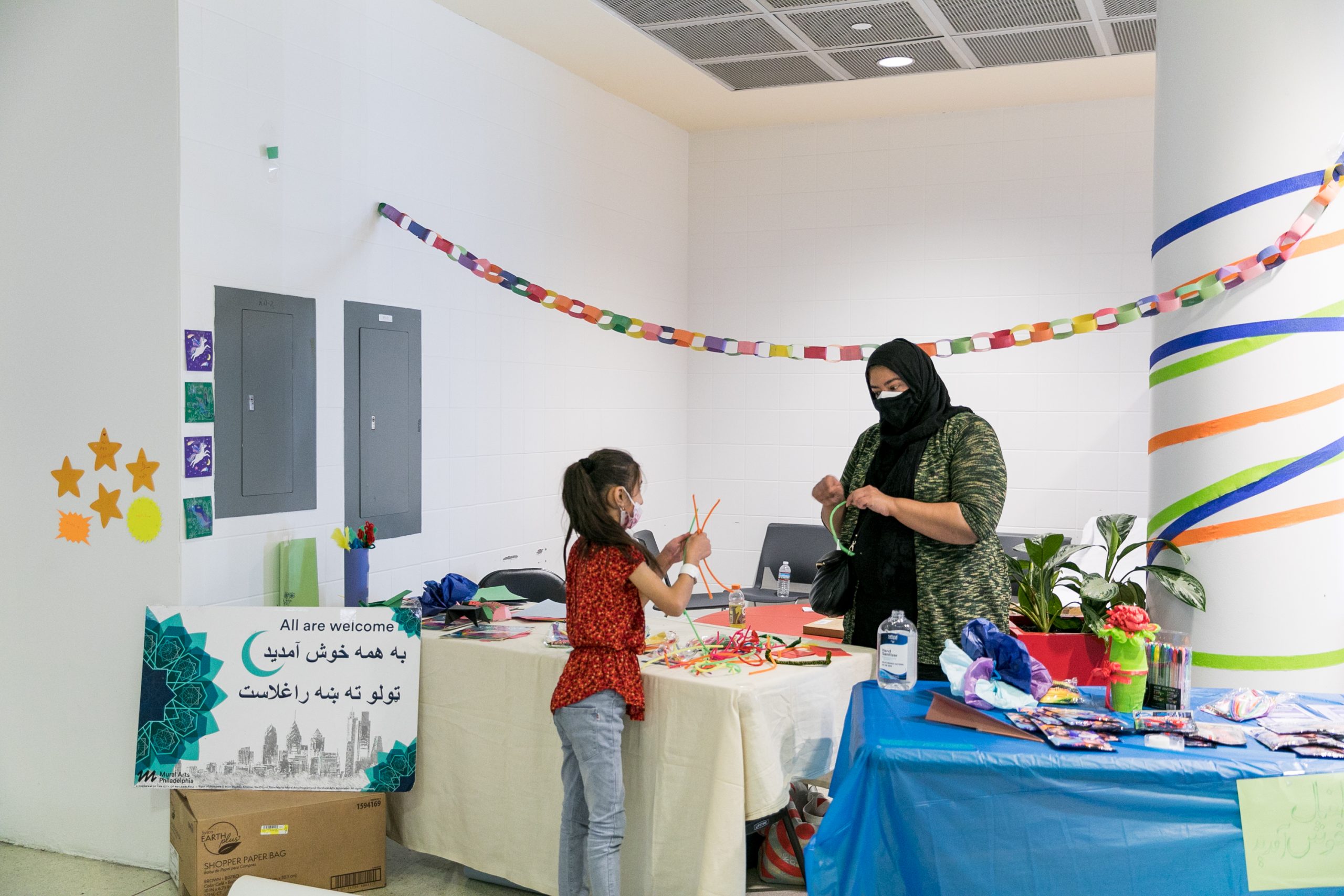 A Porch Light staff member behind a table at the airport helps a young girl create crafts. The table is brightly decorated with streamers in different colors and nearby a sign reads 
